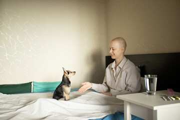 Adult female cancer patient in pajamas resting in bed with tiny dog, glass of water and pills on bedside table, home treatment