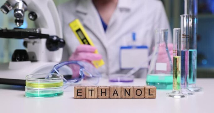 Woman lab worker measures chemical properties of sample. Wooden cubes arranged to form word Ethanol indicate focus of ongoing experiment by microscope