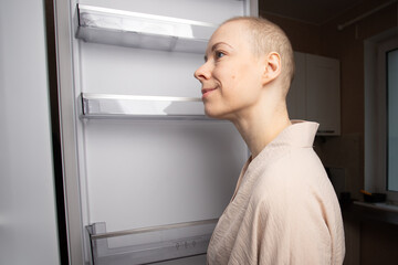 bald woman in a light bathrobe closely examines the empty shelves of an open refrigerator in her small kitchen, symbol of food choices, diet planning and healthy recovery after cancer