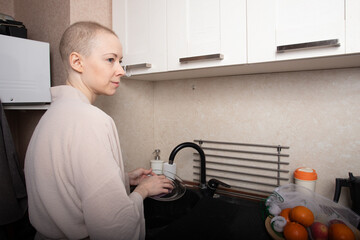 bald woman in a light bathrobe carefully washes dishes at the kitchen sink in her small apartment, symbol of daily routine, hygiene and gentle recovery after chemotherapy and serious illness