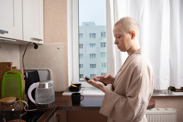 bald woman in a light bathrobe stands in front of kitchen cabinets focused on her smartphone, checking messages and health apps as part of an organized daily routine during cancer recovery