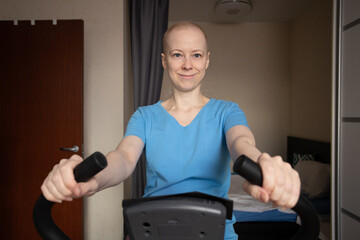 Cancer patient works out at home on an exercise bike, woman after chemotherapy training in bedroom, indoor cardio workout for oncology rehabilitation, home fitness and recovery