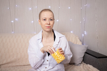 young woman with short regrowing hair after chemotherapy sits on sofa in pajamas, holding a clear plastic cup of popcorn and watching movie at home with gentle string lights in background
