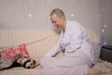 chemo patient in soft pyjamas laughing while playing with her little dog on the couch, concept of emotional support animal, mental health, cancer recovery and everyday life at home