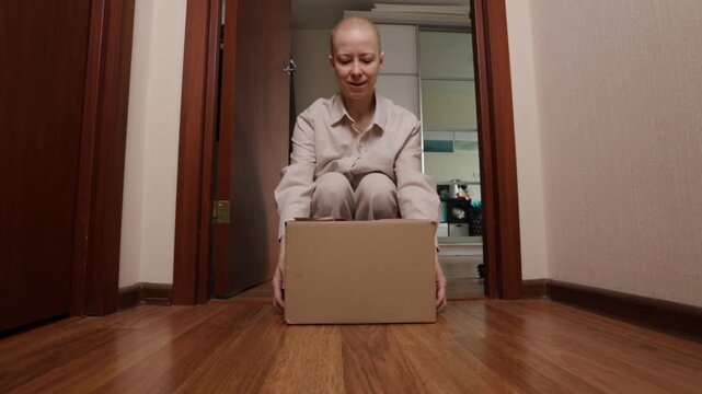 Smiling bald woman with cancer unpacking delivery box in apartment corridor, patient in loungewear receiving care package from friends, support, gifts
