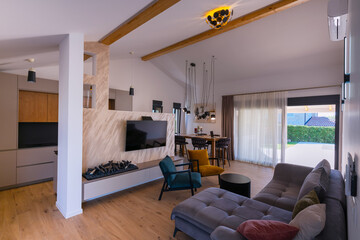 Obraz premium Modern open-plan living and dining area with an integrated kitchen, featuring a comfortable sofa, stylish armchairs, and a bar-height dining table. Wooden floors and ceiling beams enhance this