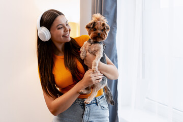 Happy woman holding her dog, enjoying music with headphones at home