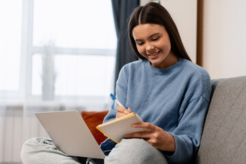 Young woman student learning, taking notes from online course on laptop at home