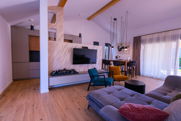 Obraz premium Modern open-plan living and dining area with an integrated kitchen, featuring a comfortable sofa, stylish armchairs, and a bar-height dining table. Wooden floors and ceiling beams enhance this