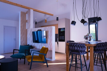 Fototapeta premium Modern open-plan living and dining area with an integrated kitchen, featuring a comfortable sofa, stylish armchairs, and a bar-height dining table. Wooden floors and ceiling beams enhance this