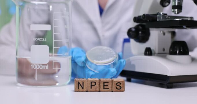Wooden blocks on lab bench spell abbreviation NPES near large beaker. Scientist in blue gloves observes sample through microscope recording result