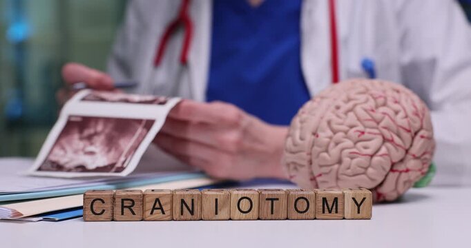 Wooden blocks on medical desk spell word Craniotomy in front of brain model. Woman doctor studies printed scans describing planned skull surgery