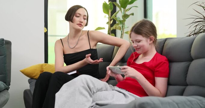 Mother sits close to daughter playing fancy game on smartphone. Angry mom scolds naughty girl about time limits while trying to set screen time rules