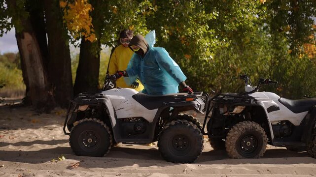Two white men with ATVs outdoors, bright yellow and teal jackets beside treelined sandy trail, one helping other start quad as dust kicks up, playful teamwork