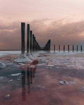 A row of old wooden posts, covered in salt crystals, extending into a pink salt lake at sunset. Conceptualizing natural beauty, salt deposits, and unusual landscapes.