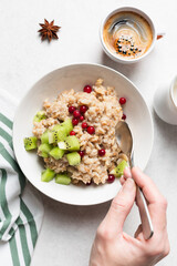 Healthy breakfast concept: woman hands holding bowl of oatmeal porridge with kiwi, cranberries and cup of coffee on side. Top view. Diet meal