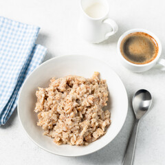 Healthy vegan breakfast meal: cooked oatmeal and spelt porridge in white bowl served with cup of coffee and almond milk in jug
