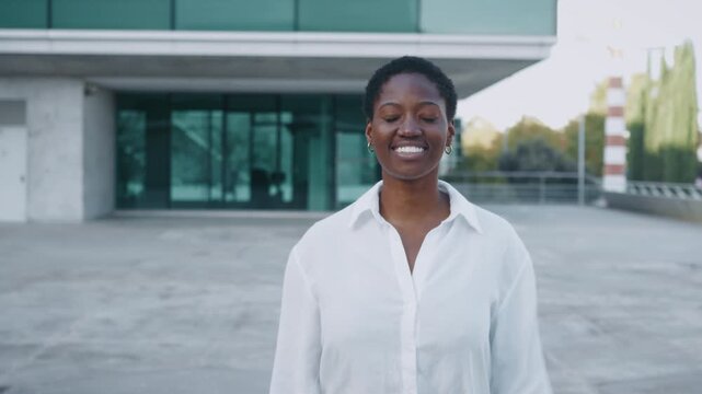 Smiling young Black businesswoman walking outside modern office building, looking at camera with confident friendly expression during daytime