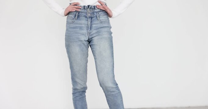 Woman in jeans places hands on hips and swings leg against white background. Female tries to relax stiff muscles before training. Slow motion