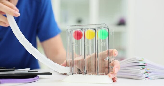 Medical worker in blue uniform blows into respiratory training device with colored balls. Doctor checks lungs capacity at desk with patients histories