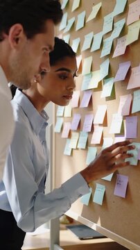 Creative professionals lean over a planning board covered with colorful sticky notes, exchanging ideas and gestures in a focused collaborative work session under soft office light.