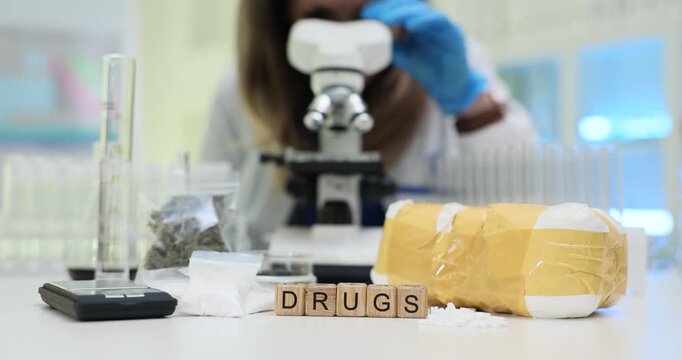 Wooden cubes spell word Drugs in front of powder wrapped package and lab tools. Forensic scientist in gloves examines evidence under microscope