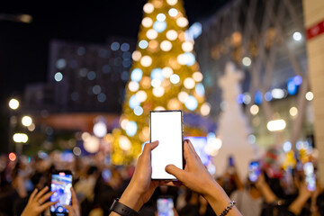 Hands Holding Smartphone with White Screen Taking Photo of Giant Christmas Tree Lights in City at Night