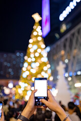 Hands Filming Giant Christmas Tree with Smartphone White Screen in City Holiday Crowd