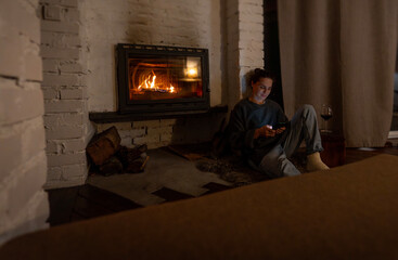 Young woman by the fireplace in a cozy house with a mobile phone in her hands