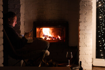 Young woman by the fireplace in a cozy house with a mobile phone in her hands
