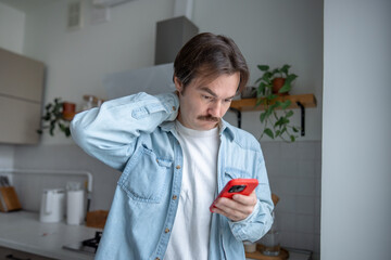 Thoughtful man standing in kitchen checking smartphone, touching neck in tension. Pensive confused male reads unexpected message on phone, feeling uncertainty, mental overload, emotional pause at home
