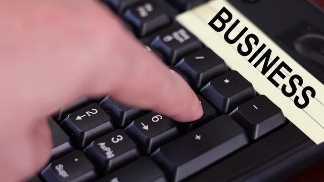 Un primer plano de un teclado con n&uacute;meros con el dedo de una mujer adulta marcando los n&uacute;meros y una nota amarilla con la palabra negocios