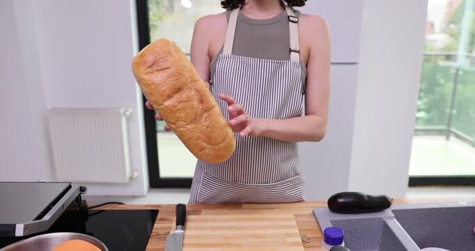 Housewife in neat apron stands at kitchen counter and carefully examines fresh loaf of bread. Female evaluates softness of bread prior to slicing