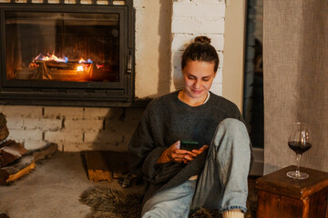 Young woman by the fireplace in a cozy house with a mobile phone in her hands