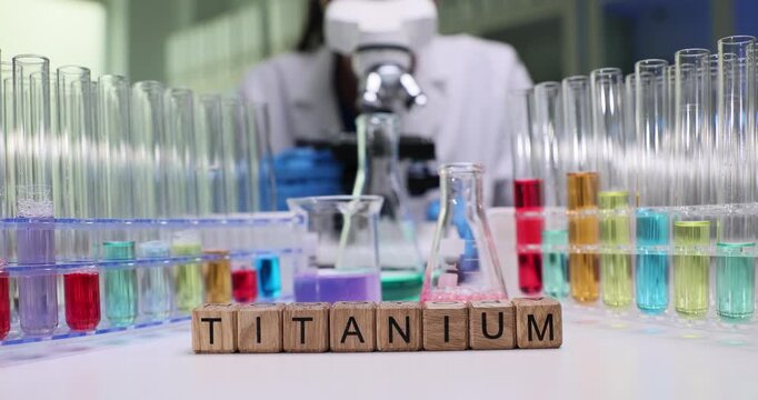 Woman chemist carefully studies structure of titanium under microscope. Wooden blocks on desk neatly form word Titanium on laboratory table with tubes