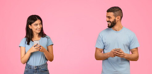 Two young adults are standing apart, engaged in a cheerful conversation while holding their smartphones. They are dressed casually and smiling at each other, creating a lively atmosphere.