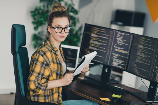 Young female programmer working in a modern office with multiple screens displaying code and notes in hand - Powered by Adobe