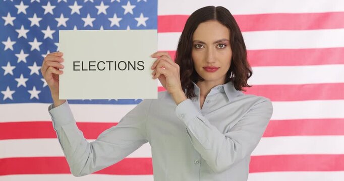 Woman stands in front of American flag holding sign with word Elections. Civic worker presents voting concept encouraging reflection on choice