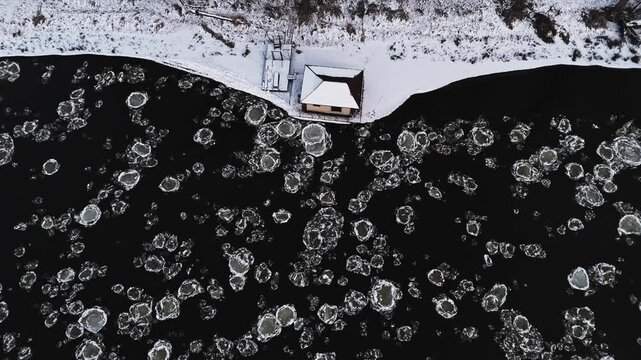 Aerial view of a dark river speckled with ice floes contrasting against a snow-covered bank and house, creating a stark winter scene, Vilnius, Lithuania.