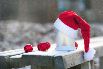 Festive lantern adorned with a red and white Santa hat, surrounded by shiny red ornaments on a snowy wooden surface, creating a cozy holiday atmosphere