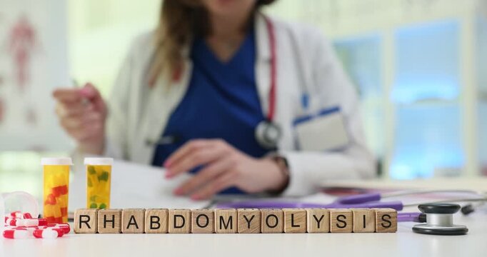 Wooden cubes spell word Rhabdomyolysis near scattered pills on table. Woman doctor writes detailed report checking serious muscle damage diagnosis