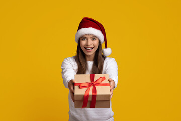 A smiling woman wearing a red Santa hat holds out a beautifully wrapped gift with a ribbon. The vibrant yellow background adds to the festive spirit, creating a cheerful holiday scene.
