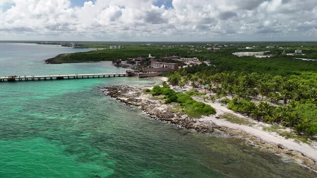 Aerial view of a pier stretching into turquoise waters, buildings nestled amidst verdant trees and a sandy beach, Mahahual, Quintana Roo, Mexico.