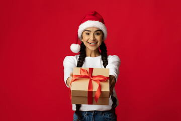 A cheerful woman wearing a Santa hat stands against a bright red background, happily presenting a wrapped gift with a red ribbon. The atmosphere is festive and joyful.