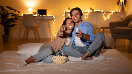 Obraz premium Millennial couple is having fun watching a movie in their bedroom. They are relaxed and pointing at the TV screen while sitting on a comfortable bed with a bowl of popcorn.