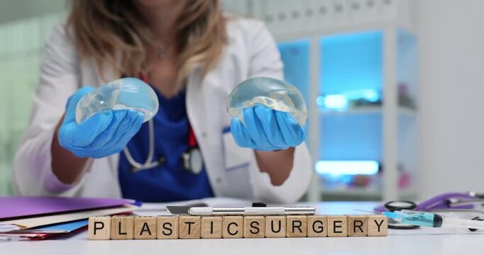 Wooden cubes spell words Plastic Surgery highlighting cosmetic procedure theme. Woman doctor wears blue gloves holding modern breast implants