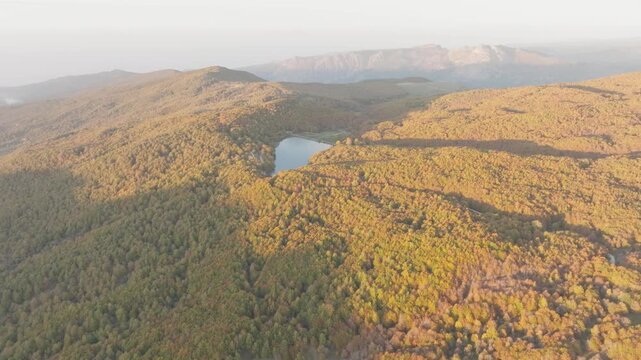 Aerial view of Lago Maulazzo; the lake's dark waters contrast with the surrounding golden forest under the hazy sky, Lago Maulazzo, Sicilia, Italy.