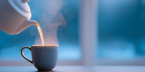 Steaming hot tea being poured from a white teapot into a dark ceramic cup on a table with a blurred blue background