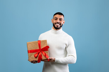 A man is smiling while holding a nicely wrapped gift with a red ribbon. He wears a cozy white...