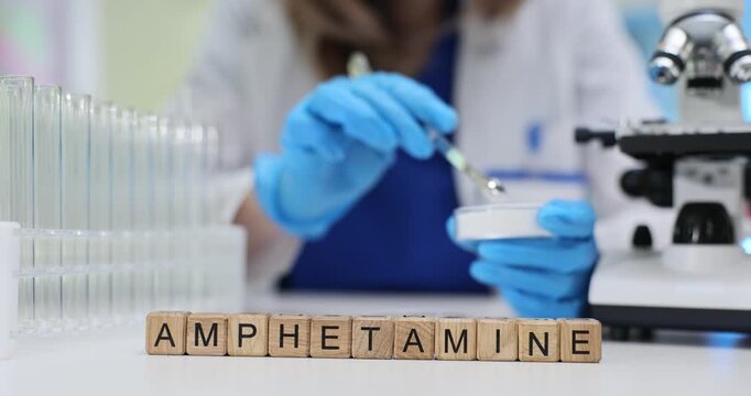Wooden blocks spell word Amphetamine near test tubes and Petri dishes. Woman scientist transfers sample drop studying illegal stimulant trace